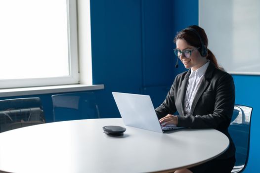 Friendly woman wearing a headset is having an online conversation on a laptop in a conference room. Female manager communicates with clients via video call. Remote business negotiations in quarantine