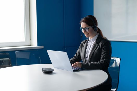 Friendly woman wearing a headset is having an online conversation on a laptop in a conference room. Female manager communicates with clients via video call. Remote business negotiations in quarantine