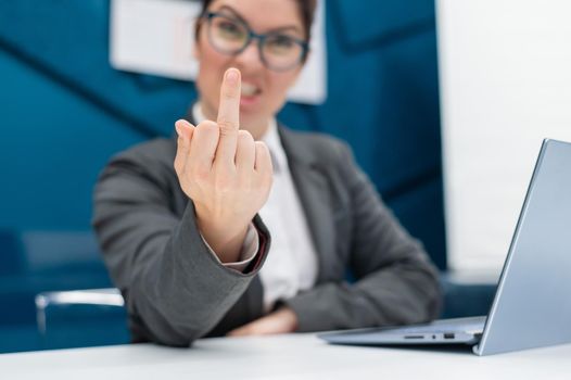 Angry business woman in a suit shows a middle finger while sitting at a desk. Annoyed female office worker showing a fuck you gesture. Professional burnout