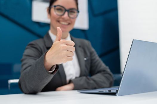 Smiling woman in a business suit shows thumb up while sitting at a desk. Female boss works at a laptop and gestures approval