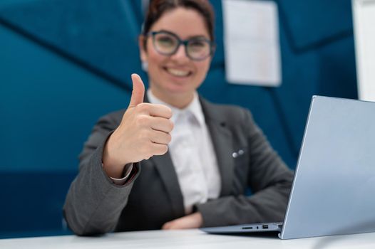 Smiling woman in a business suit shows thumb up while sitting at a desk. Female boss works at a laptop and gestures approval