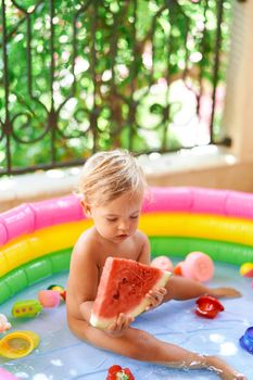 Little girl sits in an inflatable pool on the balcony with a piece of watermelon in her hands. High quality photo