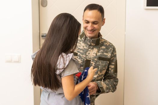 Portrait of happy american family father in military uniform and cute little girl daughter with flag of United States hugging and smiling at camera, male soldier dad reunited with family at home