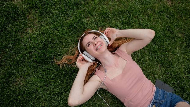 A young red-haired woman lies on the grass and listens to music