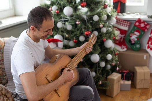 Happy young man is playing guitar. Guy is looking happily and carefree. Male in festive hat alone celebrating Christmas or new year. Christmas tree with garland in background.