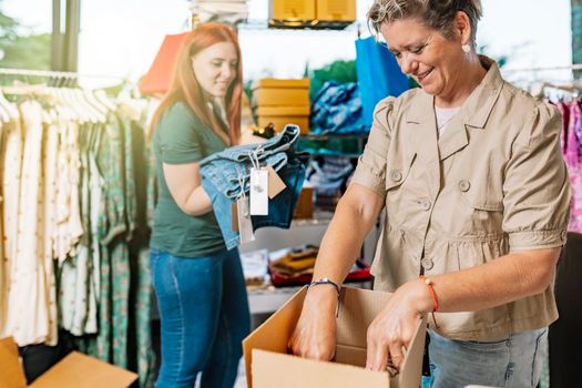 female co-workers, working in the clothing shop warehouse, preparing orders for their customers. mature female business owner and young female employee business concept and entrepreneur. natural light outdoors.