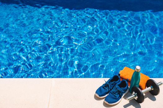 top view of sports objects in front of a swimming pool. orange towel, blue water bottle, weights. bottom of a swimming pool in a garden. outside with natural light.
