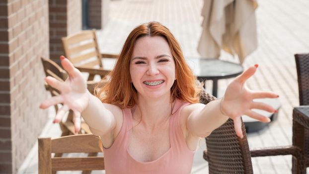 Young red-haired woman sits in a street cafe and stretches her arms to hug