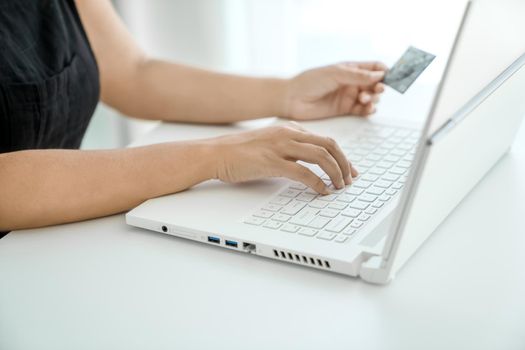 Young woman makes online purchases sitting in front of laptop with bank card in her hand. Hands close-up. Concept of online shopping and money transfer.