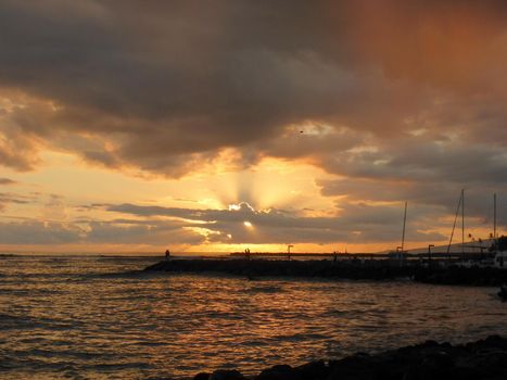 Sunsets through the clouds over the Pacific  Ocean with people watching on rock jetty with light reflecting on ocean and illuminating the sky with boats sailing on the water off Waikiki on Oahu, Hawaii.