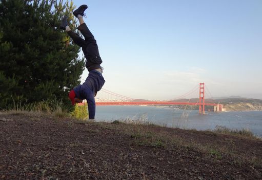 Man wearing hat, hoodie, long pants and shoes Handstands in on hill in front of the Golden Gate Bridge on the Marin side with San Francisco, California in the distance.