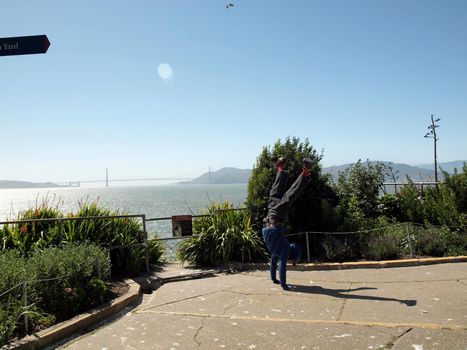 Man wearing hat, hoodie, long pants and shoes Split leg Handstand on Alcatraz Island in front of the Golden Gate Bridge with San Francisco, California in the distance.