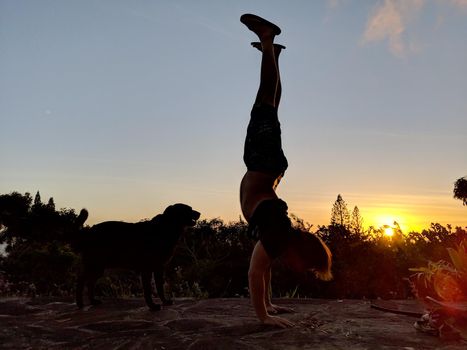 Man wearing a t-shirt, shorts, and slippers Handstands at Tantalus Mountain during sunset next to dog with lush vegetation above the City of Honolulu on Oahu, Hawaii         