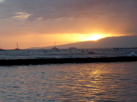 Sunsets behind Waianae mountains with light reflecting on ocean and illuminating the sky with surfers  and boats in the water off Waikiki on Oahu, Hawaii.