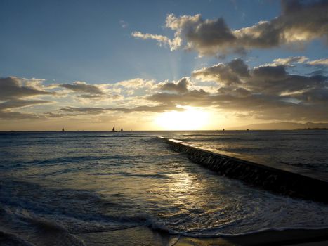 Rock Pier leads to Sunset over the Pacific ocean on the water with birds flying in the foreground and boats off the coast of Oahu, Hawaii with Waianae Mountain range visible. February 2016.