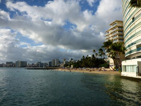 People play in the wavy water on ocean off Kaimana Beach with hotels and condos of Waikiki in distance on Oahu, Hawaii.
