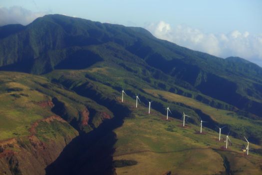 Aerial view of Modern Windmills spin on hillside with windy road up to mountain top on the island of Maui.