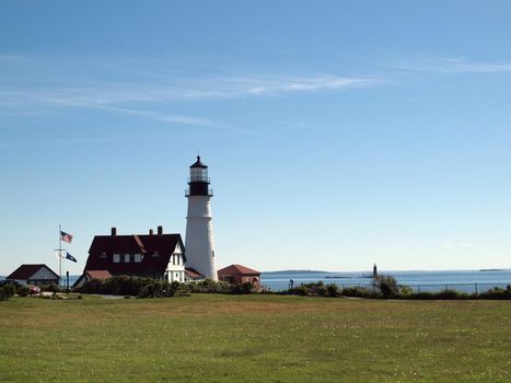 Portland Head Lighthouse and Keepers' Quarters building with lighthouse on island in distance in Portland, Maine.  Portland Head Light, is a historic lighthouse in Cape Elizabeth, Maine. The light station sits on a head of land at the entrance of the primary shipping channel into Portland Harbor, which is within Casco Bay in the Gulf of Maine. Completed in 1791, it is the oldest lighthouse in the state of Maine. The light station is automated, and the tower, beacon, and foghorn are maintained by the United States Coast Guard, while the former lighthouse keepers' house is a maritime museum within Fort Williams Park. Keepers' Quarters building was constructed in 1891 as a two story duplex. Until 1989, it was home to the head and assistant lighthouse keepers and their families.