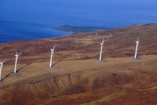 Aerial view of Modern Windmills spin on hillside with windy road and coastline on the island of Maui.