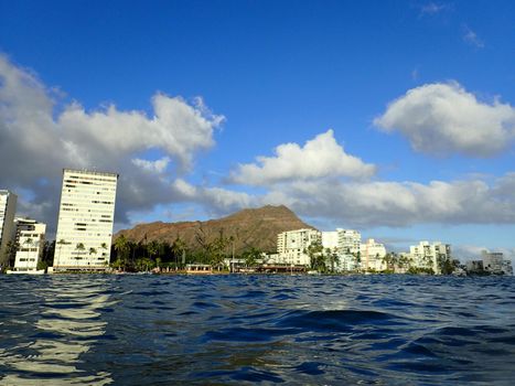 Hotel building, Outrigger Canoe Club, coconut trees, Condo buildings, clouds, and Diamond Head Crater in the distance on Oahu, Hawaii viewed from the water on a beautiful day. 