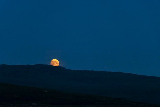 Bright orange worm super moon with patterns and craters visible, partially obscured as it rises over the incline of a mountain in a clear, dark blue sky.