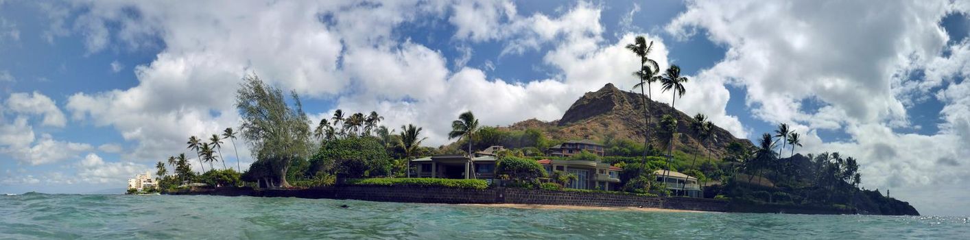 Panoramic of Beach with waves lapping, napakaa, lava rock wall and Coconut trees with Diamond Head in the background along the shore on a wonderful day in Oahu, Hawaii.