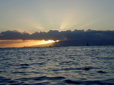 Sunsets through the clouds over Waianae mountains with light reflecting on ocean and illuminating the sky with boats sailing on the water off Waikiki on Oahu, Hawaii.