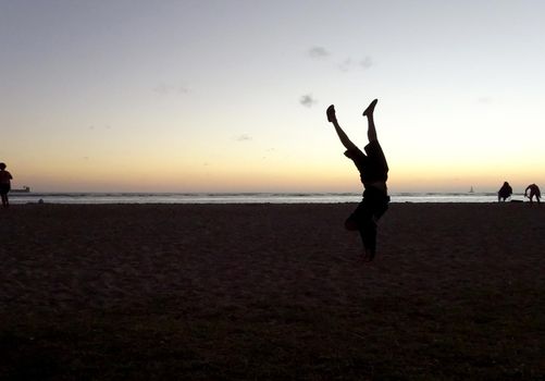 Man Handstanding on beach at sunset with boats on the ocean at Magic Island on Oahu, Hawaii.                    