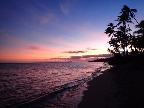 Sandy shoreline of Kahala Beach and the southern coastline at dusk with pink and orange light filling the air of Oahu, Hawaii.