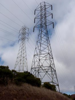 High Voltage Power Lines intersect at two large metal Utility pole with bushes on the ground against a blue sky in California.