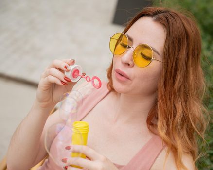 Young red-haired woman blowing soap bubbles outdoors. Girl in yellow sunglasses and braces