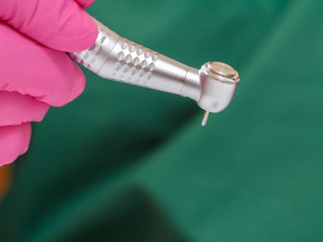 Dentist's hand in a pink latex glove with a new high-speed dental handpiece and blurred green medical gown on the background. Medical tools concept.