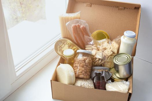 Grocery set of foodstaff for needy in crisis, pasta, beans, canned food, oil, milk, sausage, cheese in cardboard box on the windowsill, food donation concept, top view