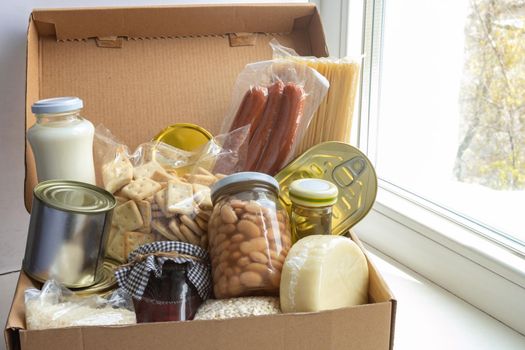 Grocery set of foodstaff for needy in crisis, pasta, beans, canned food, oil, milk, sausage, cheese in cardboard box on the windowsill, food donation concept