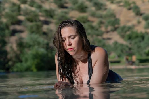 attractive young long-haired brunette girl in black swimsuit lying on the shore of a lake looking at the water