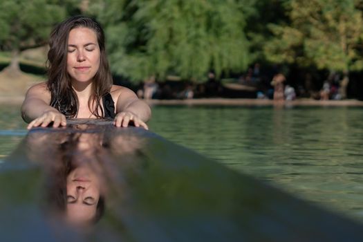 attractive young latin girl reflected in the water of a lake with eyes closed leaning on a wooden board