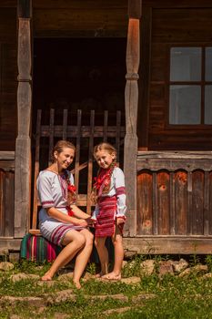 mother and daughter in Ukrainian national costumes are sitting near an old house