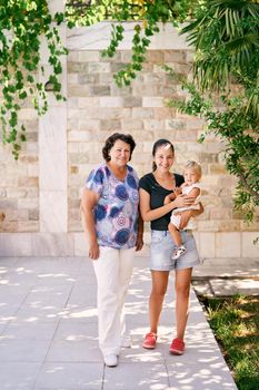 Grandmother stands next to mother holding a little girl in her arms in a garden near a stone wall. High quality photo