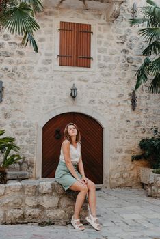 Girl Tourist Resting in the Ancient Narrow Street On A Beautiful Summer Day In MEDITERRANEAN MEDIEVAL CITY, OLD TOWN KOTOR, MONTENEGRO. Young Beautiful Cheerful Woman Walking On Old Street