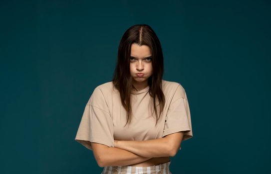 Portrait of upset young beautifil girl in a beige t-shirt standing with puffy cheeks and arms folded isolated over blue background. Emotion of angry and upset