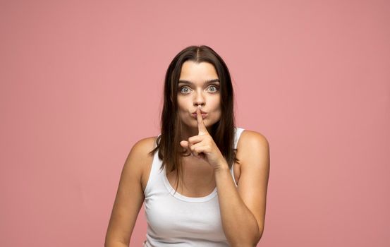 Emotional beautiful brunette woman with attractive appearance, keeps fore finger on lips, demonstrates silence gesture and ask to keep confidential information in secret. Pink studio background