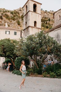 Girl Tourist Resting in the Ancient Narrow Street On A Beautiful Summer Day In MEDITERRANEAN MEDIEVAL CITY, OLD TOWN KOTOR, MONTENEGRO. Young Beautiful Cheerful Woman Walking On Old Street