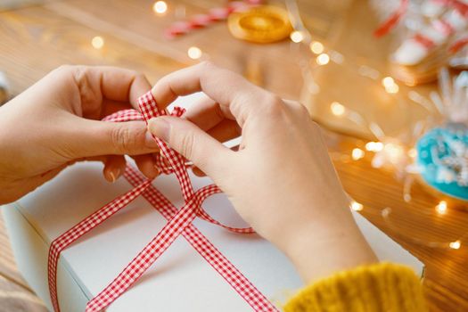 Person's hands in knitted sweater tie red ribbon bow on white gift box. Garland lights, gingerbread and decorations in background. New Year's mood. Surprise for Birthday or Christmas.