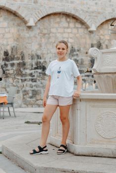 Girl Tourist Resting in the Ancient Narrow Street On A Beautiful Summer Day In MEDITERRANEAN MEDIEVAL CITY, OLD TOWN KOTOR, MONTENEGRO. Young Beautiful Cheerful Woman Walking On Old Street