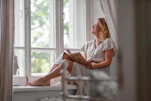 A middle-aged woman in a cream dress sits mysteriously and looks out the window on the windowsill. Green trees outside