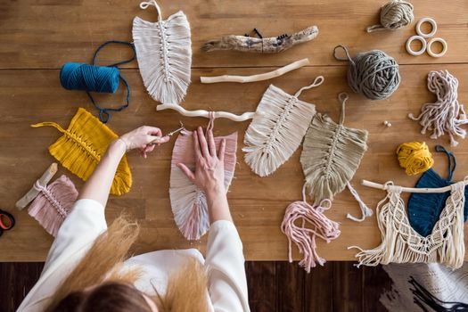 Small home business concept. Top view of woman creating macrame on wooden table. Female hands cutting macrame feathers or leaves with scissors