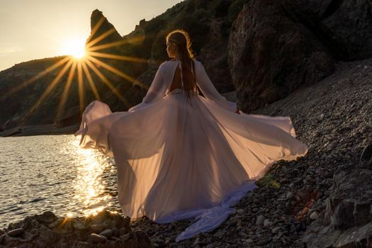 Mysterious woman silhouette long hair walks on the beach ocean water, sea nymph wind listens to the wave. Throws up a long white dress, a divine sunset. Artistic photo from the back without a face.