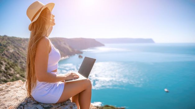 Successful business woman in yellow hat working on laptop by the sea. Pretty lady typing on computer at summer day outdoors. Freelance, travel and holidays concept.