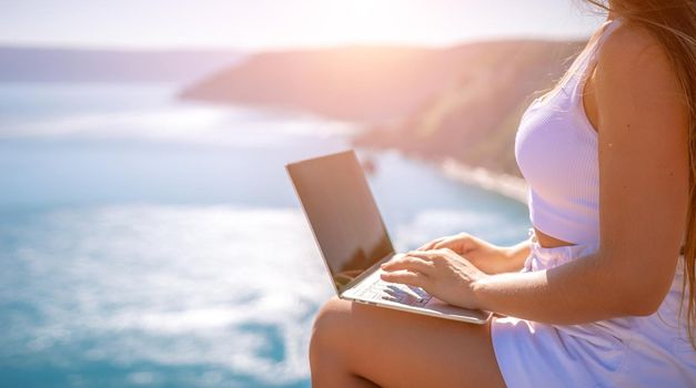 Successful business woman in yellow hat working on laptop by the sea. Pretty lady typing on computer at summer day outdoors. Freelance, travel and holidays concept.