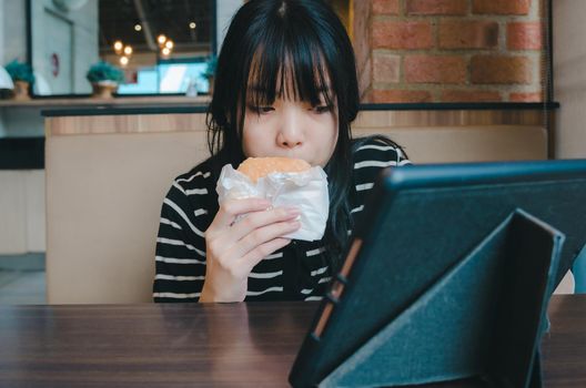 young girl is eating a hamburger and watching an online social media tech tablet on the table.